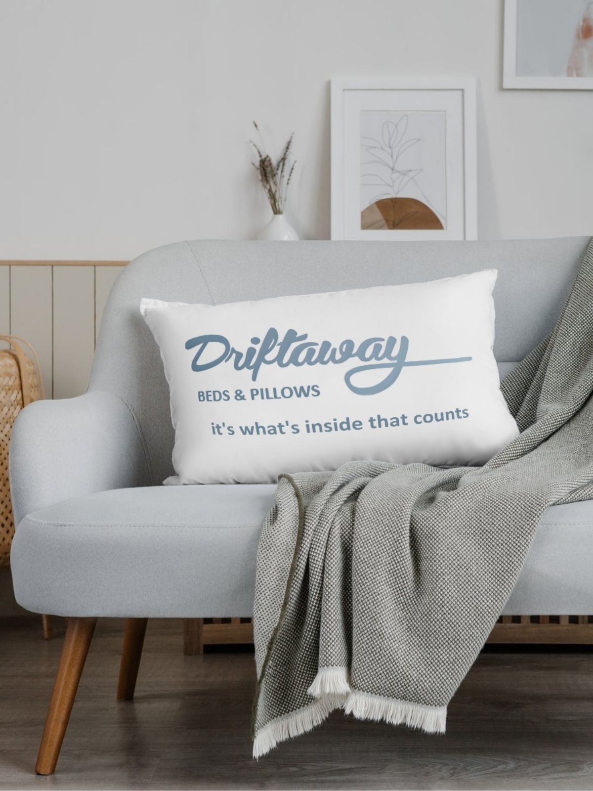 White pillow with 'Driftaway Beds & Pillows' text on a gray sofa with a blanket.